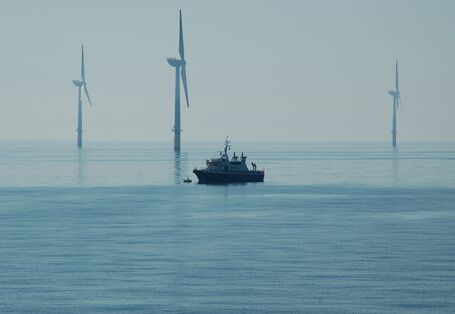 Das Foto zeigt ein Schiff vor drei Windenergieanlagen mit einem Monopile-Fundament. Davor sieht man ein im Verhältnis sehr klein wirkendes Schiff.