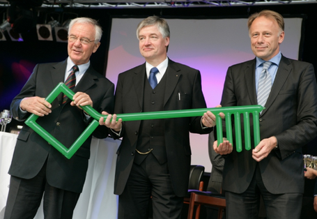 Gruppenfoto: drei Männer in Anzug und Krawatte halten einen riesigen grünen Schlüssel, im Hintergrund Sektgläser