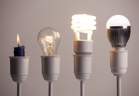 vier Lampenfassungen in einer Reihe, darin stecken (von links nach rechts): eine Kerze, eine Glühlampe, eine Energiesparlampe (Kompaktleuchtstofflampe), eine LED-Lampe