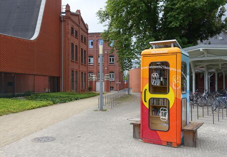 eine bunt angemalte Telefonzelle ist innen zu einer Miniblibliothek ausgebaut, auf dem Dach ein Solarmodul, außen eine Holzbank