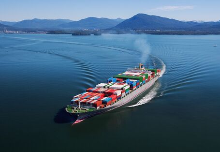 Foto: Containerschiff von oben, das über das Meer fährt. Im Hintergrund Berge. Eine Abgasfahne steigt aus dem Schornstein.