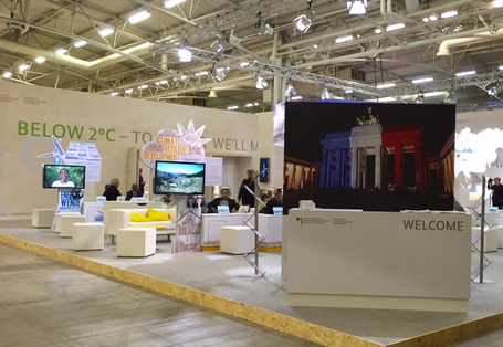 in einer Messehalle sind Stellwände, Sitzgelegenheiten und Bildschirme aufgebaut. Eine Stellwand zeigt das Brandenburger Tor, das in den Farben der französischen Flagge angestrahlt ist. Auf einer Stellwand hinten steht "Below 2 °C - together we´ll make it