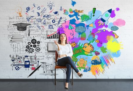 eine Frau sitzt vor einer Wand, im Hintergrund ist eine Hälfte der Wand in schwarz-weiß mit Symbolen für Wissen bemalt, die andere in bunt mit Symbolen für Kreativität