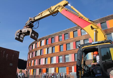 UBA-Präsident Flasbarth sitzt vor dem Dienstgebäude in Dessau-Roßlau in einem Bagger, um eine alte Wand aus Ziegeln einzureißen, Menschen am Bauzaun schauen zu