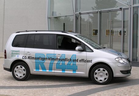 ein silbergrauer VW Touran steht vor dem UBA Dessau-Roßlau. Er trägt die Beschriftung "CO2-Klimaanlagen - klimafreundlich und effizient, R744" und das Logo des Umweltbundesamtes