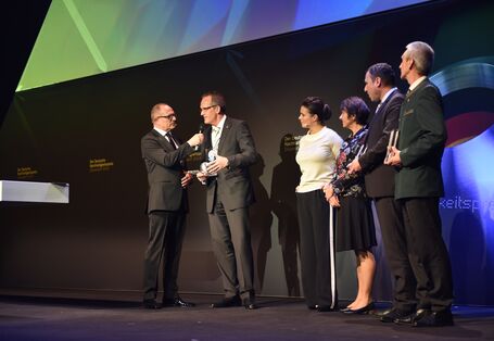auf einer Bühne: ein Mann im Anzug hält eine Blauer Engel-Trophäe in der Hand und wird mit einem Mikrofon befragt, daneben in einer Reihe weitere Personen