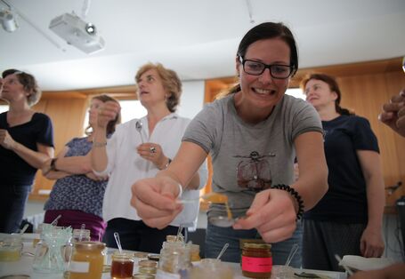 eine Gruppe Frauen vor einem Tisch mit Honiggläsern, eine junge Frau entnimmt Honig mit einem Teelöffel