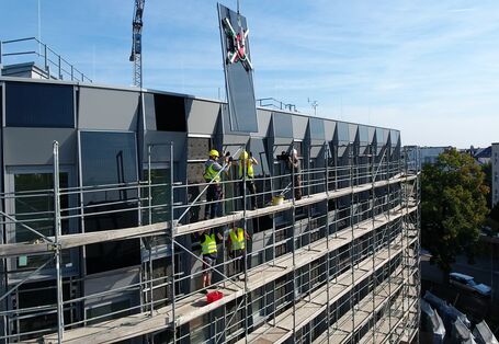eingerüstetes Gebäude mit Fassade aus Photovoltaikmodulen, Bauarbeiter auf dem Gerüst nehmen ein Modul in Empfang, welches an einem Kran hängt