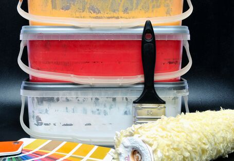 verschiedenfarbige Wandfarben in niedrigen Eimern, davor ein Fächer mit Farbmuster-Bögen, ein Pinsel und eine Rolle zum Wändestreichen