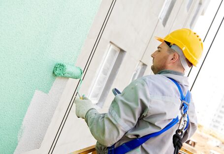 Man in overalls and yellow hard hat applying a mint green coat to a building façade