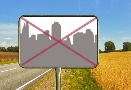 An einer Landstraße, die durch Felder führt, steht ein Ortsschild. Per Fotomontage ist in das Schild eine Stadtsilhouette mit vielen Gebäuden gemalt und diese ist rot durchgestrichen.