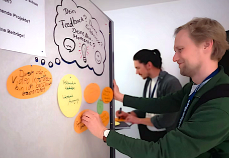 zwei Männer bringen Zettel an einer Stellwand zum Thema "Dein Feedback" an