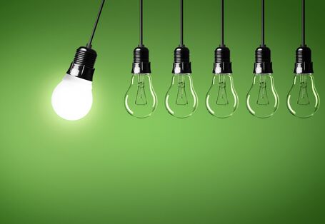 vor grünem Hintergrund schwingt eine leuchtende Energiesparlampe in einer Lampenfassung am Kabel gegen eine Reihe nicht leuchtender Glühlampen