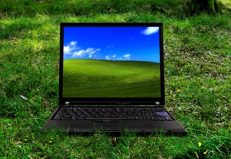 ein Laptop steht auf einer grünen Wiese, auf dem Bildschirm ist ein grüner, grasbewachsener Hügel vor blauem Himmel zu sehen
