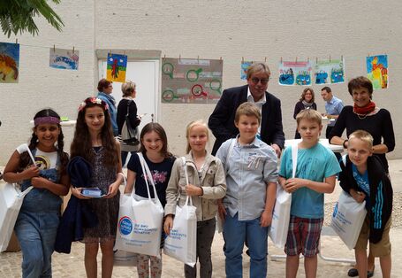 vier Mädchen und drei Jungen stehen in einer Reihe lächelnd vor der Kamera mit gefüllten Baumwollbeuteln mit der Aufschrift "Die BMU Klimaschutzinitiative". Dahinter Dr. Thomas Holzmann und Maria Krautzberger und die Kinderbilder auf der Wäscheleine.