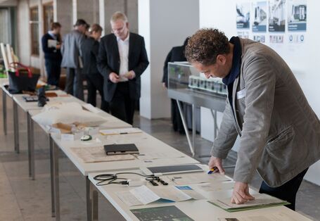 Auif einer langen Tischreihe liegen Produkte und Ausdrucke, die von mehreren Personen angeschaut werden