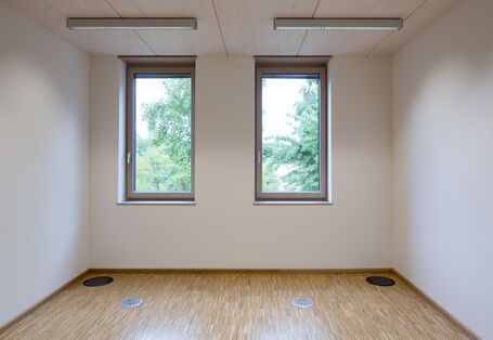 leerer Büroraum mit weißen Wänden und Holzfußboden, durch die zwei Fenster sind grüne Baumkronen zu sehen