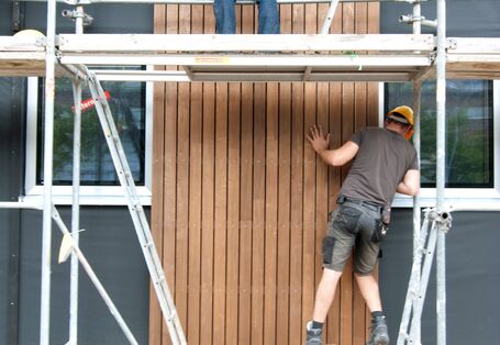 zwei Bauarbeiter stehen auf einem Baugerüst und montieren zusammen ein Holz-Fassadenelement