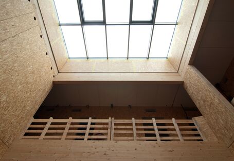 Innenansicht des Gebäudes im Rohbauzustand, die Innenwände aus rohen Holzwerkstoffplatten, durch ein Glasdacht fällt Tageslicht durch das Treppenhaus bis ins Erdgeschoss