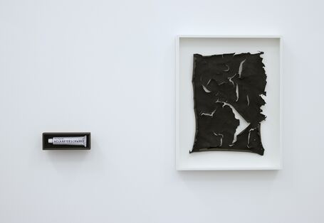 an einer weißen Wand hängt eingerahmt ein kohlrabenschwarzes, zerrissenes Blatt Papier oder Haut, daneben hängt, schwarz eingerahmt, eine Tube mit der Aufschrift "Neckartorschwarz" 