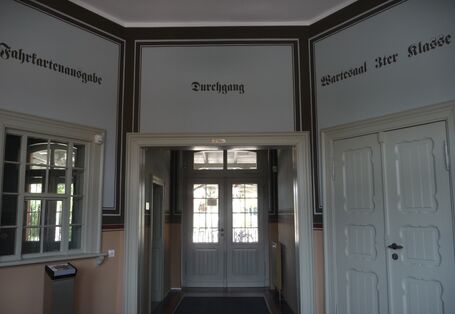 Gebäudeinnere, links eine alte Fahrkartenausgabe mit hochschiebbarer Glasscheibe, über einer Holz-Doppeltür steht in kunstvoll aufgemalten Buchstaben "Wartesaal 3ter Klasse".
