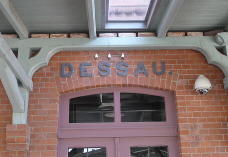 Eingangstür eines Backsteingebäude über der "Dessau" steht, darüber ein Vordach aus grau gestrichenem Holz
