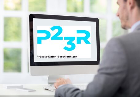 ein junger Mann im Anzug sitzt im Büro an einem Schreibtisch. Auf dem Computermonitor steht "P23R Prozess-Daten-Beschleuniger"