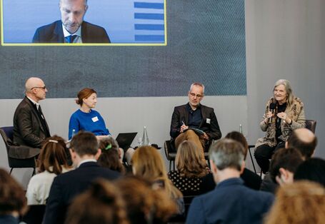 Vier Leute sitzen auf Stühlen auf einer Bühne und reden miteinander, man sieht die Rücken der im Publikum sitzenden Teilnehmer*innen.