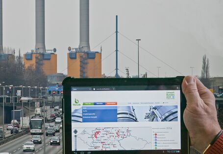 eine Hand hält ein Tablet mit dem Webangebot thru.de vor ein Kraftwerk hinter einer Stadtautobahn