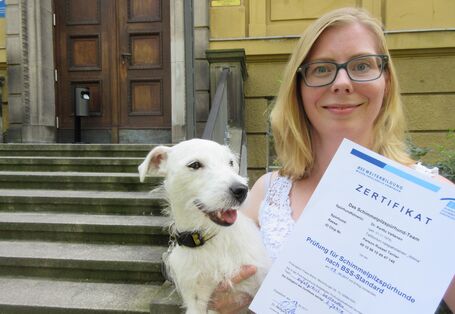 eine Frau hält mit der einen Hand einen weißen Terrier und mit der anderen ein Blatt Papier mit der Aufschrift "Zertifikat"