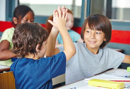 zwei Grundschüler geben sich, im Klassenzimmer an ihrem Tisch sitzend,  einen Handschlag. Im Hintergrund sitzen zwei Schülerinnen an ihrem Tisch. Auf den Tischen liegen Papier und Stifte.