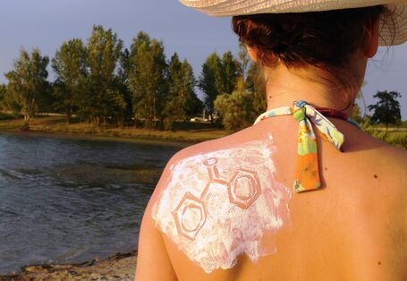 Eine Frau mit Sonnenhut und Bikini steht an einem See. Man sieht Rücken und Hinterkopf. Das linke Schulterblatt ist mit Sonnencreme eingecremt, in die eine chemische Strukturformel gezeichnet ist.