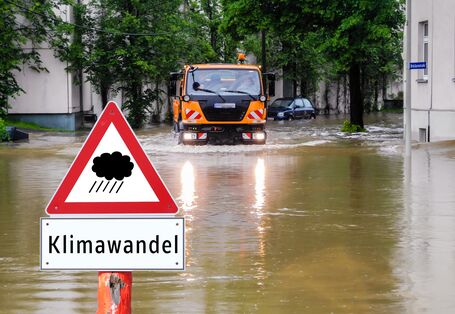 überflutete Straße in der Stadt, davor ein Warnschild "Regen / Klimawandel", ein kommunales Einsatzfahrzeug bahnt sich den Weg durchs Wasser