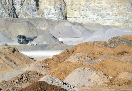 in einem Steinbruch werden Baustoffe abgebaut, man sieht verschiedenfarbige Schüttguthalden und einen Lkw vor einer steilen Felswand