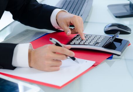 Mann im Anzug sitzt am Schreibtisch und rechnet mit einem Taschenrechner