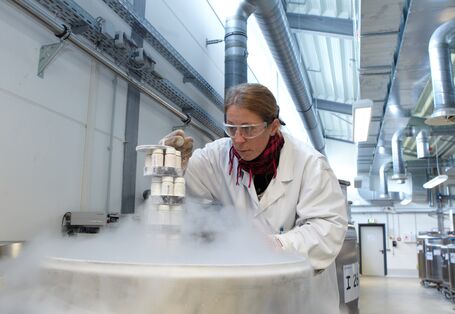 eine Frau in einem weißen Kittel, mit Schal, Handschuhen und Schutzbrille holt Proben aus einem dampfenden Metallbehälter, der in einer Halle steht