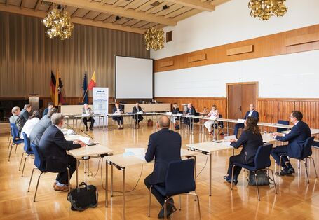 in einem Saal sitzen Männer und Frauen an Tischen im Kreis, hinten ein Aufsteller und Fahnen