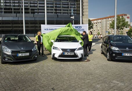 drei Autos stehen auf einem gepflasterten Platz, eine Frau und ein Mann ziehen ein grünes Tuch vom mittleren Auto, um es zu enthüllen. Im Hintergrund steht ein Banner des Verkehrsclubs Deutschland