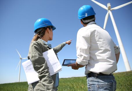eine Frau und ein Mann mit Bauhelm, Plänen und Tablet schauen sich einen Windpark an