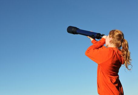 eine junge Frau schaut mit einem Fernrohr in die Ferne, im Hintergrund klarer, blauer Himmel
