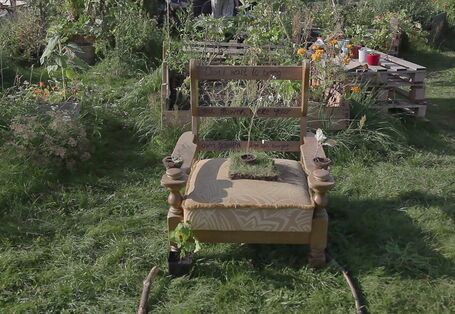 Ein Garten mit mehreren Beeten, in der Mitte steht ein alter Sessel, aus dem eine Pflanze wächst.