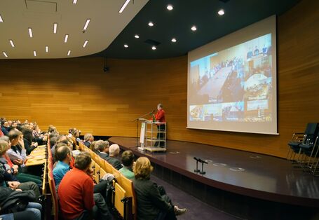 Barbara Hendricks hält im Hörsaal des UBA eine Rede, im Hintergrund sieht man auf einer Leinwand die Videoübertragung an andere Standorte. Im Vordergrund das Publikum in Dessau.