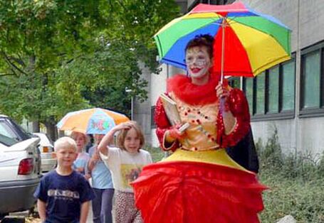 Eine Frau als Clown verkleidet hält einen bunten Regenschirm und läuft mit einigen Kindern auf einem Bürgersteig.