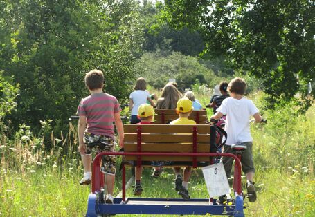 Eine Gruppe von Kindern sitzt auf einer Draisine auf Schienen und treibt sie mit Fahrradpedalen an.