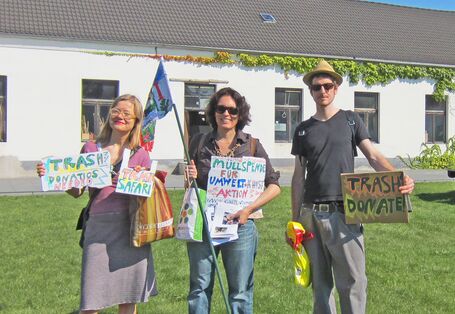 Zwei Frauen und ein Mann halten Schilder hoch, dass sie auf der Suche nach Müll sind. Sie stehen auf einer Wiese vor einem Haus.