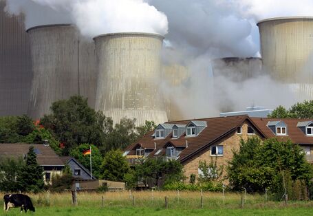Eine Kuh grast auf einer Wiese, dahinter stehen Einfamilienhäuser. Hinter den Häuser ragen riesige qualmende Schornsteine empor.