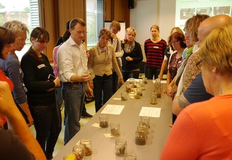 Menschen gruppieren sich um einen Tisch, auf dem Gläser mit verschiedenen Materialien stehen und die erläutert werden