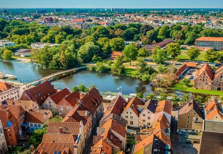 Der Blick geht von oben in die Ferne über eine Stadt mit roten Ziegelsteindächern. Die Stadt ist umgeben von Grün und liegt an einem Fluss. Der Himmel ist blau.