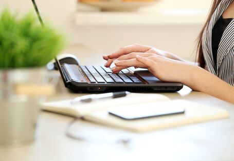 Eine Frau schreibt auf einem Notebook. Vor ihr steht ein Blumentopf mit einer Grünpflanze..