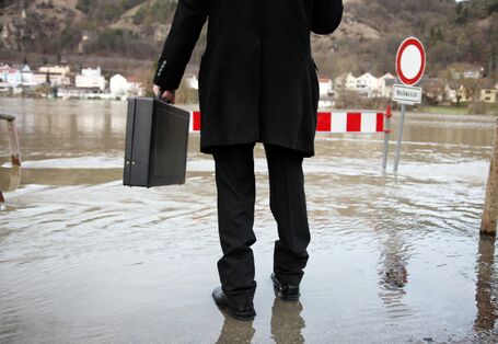 Rückenaufnahme eines Mannes im schwarzen Anzug mit Aktentasche, der auf überfluteter Fläche steht.